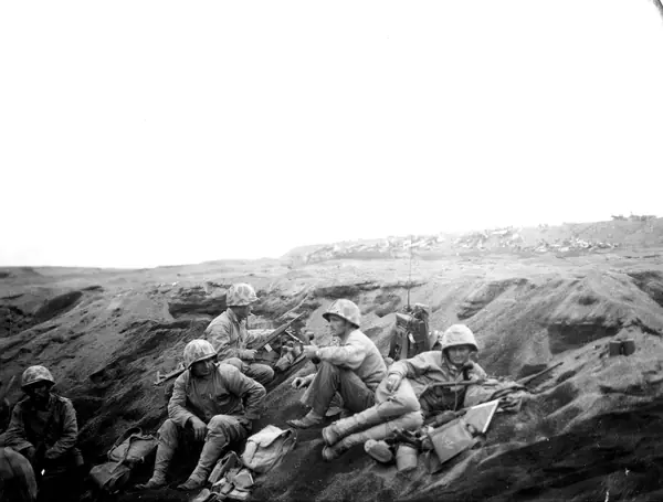 A Marine assault squad on beach at Iwo Jima awaits orders to move out. Wrecked planes on landing strip in background. (53409940181)